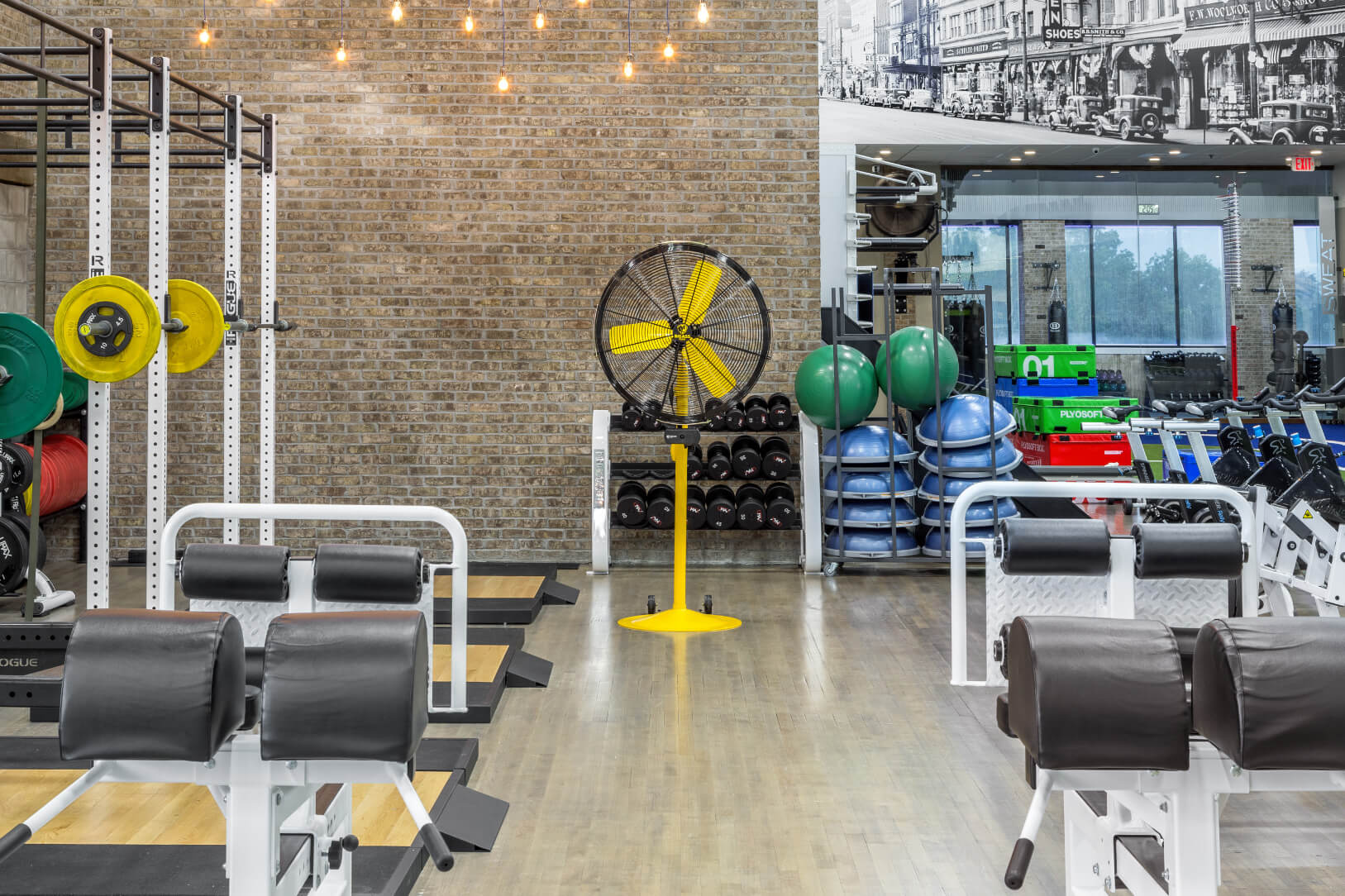 Gelber Fitnessventilator mit schwarzem Gehäuse steht auf einem Standfuß in einem Fitnessstudio